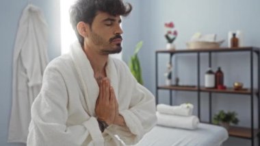 Genç adam sağlık merkezinde meditasyon yapıyor, beyaz bir bornoz giyiyor ve kaplıca dekoruyla çevrili bir yatakta oturuyor.
