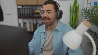 Bıyıklı, kulaklık takan ve gözlüklü genç İspanyol adam modern ofis içinde çalışıyor. Arka planda kitap rafları ve lamba var.