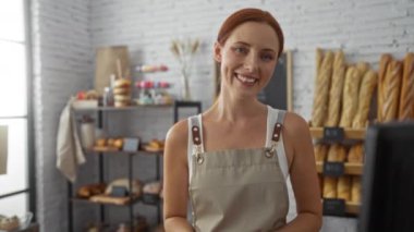 Arka planda ekmek somunları olan bir fırında gülümseyen kadın, bej bir önlük giyiyor, sıcak bir dükkanda elinde fırınlanmış mallarla bekliyor.
