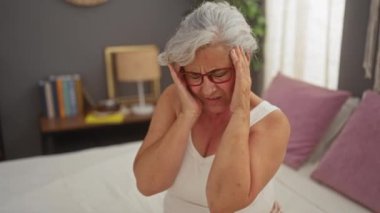 Gri saçlı yaşlı bir kadın, yatak odasında kapalı bir odada oturuyor. Başını tutuyor ve gözlüğünü çıkarıyor.
