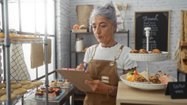 Elderly woman with grey hair takes notes on a clipboard in a cozy bakery filled with freshly baked pastries and desserts, wearing an apron and standing near shelves and a menu board. clipart