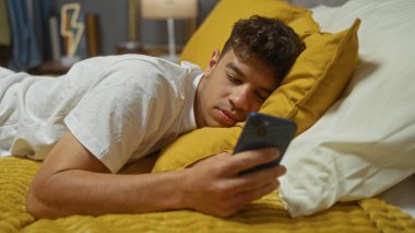 Yakışıklı, genç, İspanyol bir adam sarı bir yatakta uzanıyor, telefonunu kullanıyor, rahat ve samimi bir ortam yaratıyor..