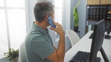 Ofiste telefonla konuşan ve arkasında bilgisayar ve bitkiler olan bir adam.