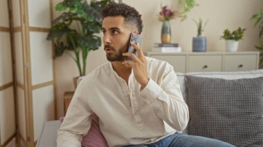 Yakışıklı, genç, İspanyol asıllı, evinin oturma odasında otururken telefonla konuşan, modern iç dekorasyonlu rahat bir ev ortamı sergileyen bir adam..