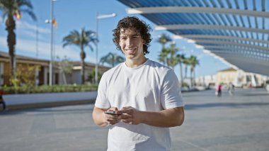 Yakışıklı, genç, kıvırcık saçlı bir İspanyol, güneşli bir caddede akıllı telefon kullanırken gülümsüyor..