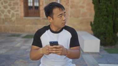 Şehir dışındaki bir şehir parkında akıllı telefon tutan genç, yakışıklı bir Çinli adam. Düşünceli ve sıradan bir tişört giymiş..