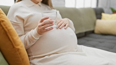 Kanepede otururken elinde bir bardak su tutan hamile bir İspanyol kadın..