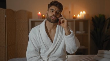 Sakallı genç İspanyol bir adam arka planda mumlarla loş bir sağlık spa odasında bornoz giyerken akıllı telefondan konuşuyor.