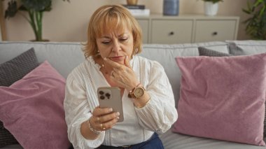Olgun, sarışın, beyaz bir kadın modern bir oturma odasında oturmuş, akıllı telefonuna dikkatlice bakıyor pembe ve gri yastıklarla süslenmiş bir kanepede dinleniyor..