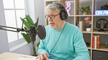 Gri saçlı, kulaklık takan olgun bir adam radyo stüdyosunda mikrofona konuşuyor..