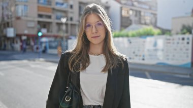 Gündelik şık giyinmiş kendine güvenen genç bir kadın güneşli bir caddede duruyor..