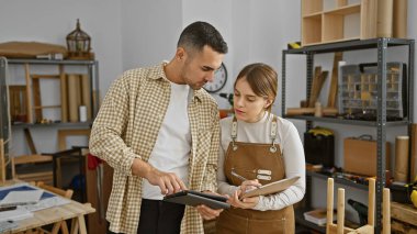 Man and woman review designs on tablet in a well-equipped woodworking workshop indoors. clipart