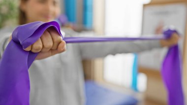Odaklanmış genç bir kadın, parlak bir fizyoterapi kliniğinde direniş bantlarıyla egzersiz yapıyor, sağlık ve rehabilitasyonu somutlaştırıyor..