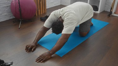 Yakışıklı Afrikalı Amerikalı bir adam kapalı spor salonundaki mavi bir paspasın üzerinde esneme egzersizi yapıyor..