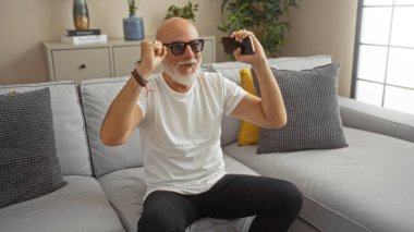 Gri saçlı ve sakallı bir adam oturma odasında bir koltukta oturuyor, bir telefon tutuyor ve kollarını kaldırarak kutluyor.