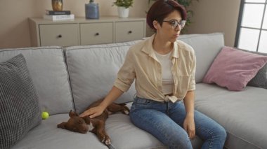 Güzel, genç, beyaz bir kadın evindeki oturma odasındaki bir kanepede oturuyor, şefkatle kahverengi bir köpeği okşuyor, yanında dinleniyor..