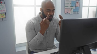 Yakışıklı, kel, Afrikalı, sakallı bir Amerikalı iş yerinde bilgisayar ekranına bakarken parmaklarını çaprazlıyor.