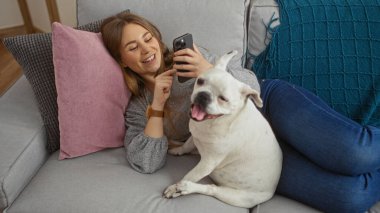 Genç sarışın bir kadın oturma odasında dinleniyor, telefonuna gülümsüyor ve evcil köpeği kanepede yanında mutlu bir şekilde oturuyor..