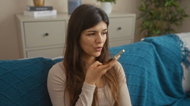 Genç ve çekici bir İspanyol esmer kadın, evinde rahat bir oturma odasında, rahat mavi battaniyeler ve modern dekorlarla otururken telefonuyla konuşuyor..