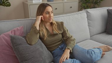 Genç, güzel, sarışın bir kadın kentsel bir dairede rahat bir koltukta dinleniyor, modern dekorlar ve yumuşak yastıklarla huzurlu bir atmosfer yaratıyor..