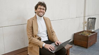 Sakallı yakışıklı İspanyol bir adam kentte dizüstü bilgisayarını kullanarak bankta oturuyor..