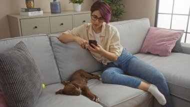 Güzel, genç bir kadın oturma odasında rahat bir şekilde telefonunu kullanırken evcil köpeği kanepede huzur içinde uyuyor..