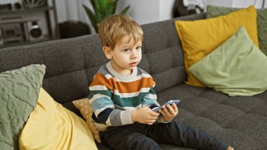 Sarışın çocuk gri bir kanepede renkli yastıklarla oturuyor, rahat bir oturma odasında akıllı bir telefona odaklanmış.