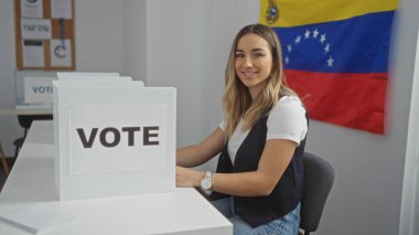 Genç, çekici, sarışın, beyaz bir kadın Venezuela 'da kapalı bir seçim salonunda oturuyor. Arka planda Venezuela bayrağı olan oy verme kabininin yanında gülümsüyor..