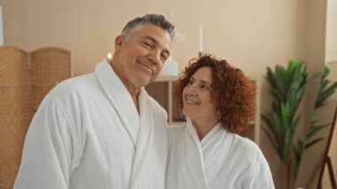 Beyaz cüppeli orta yaşlı bir çift sağlıklı yaşam kaplıcasının tadını çıkarıyorlar. Kadın adama sıcak bir şekilde gülümsüyor. Etrafı kaplıca dekoru ile çevrili..