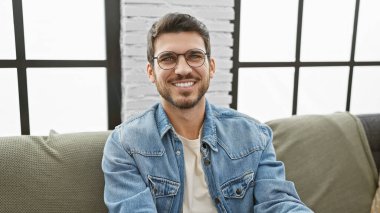 Gözlüklü ve sakallı gülümseyen genç adam, içeride kot ceket giyiyor, günlük ev tarzını tasvir ediyor..