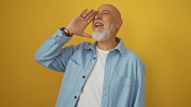 Gri saçlı ve sakallı adam gülüyor, mavi gömlek giyiyor, el kol hareketi yapıyor, sarı arka planda izole edilmiş, kendine güveni ve mutluluğu yayıyor..