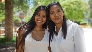 Anne ve kızı güneşli bir şehir parkında kucaklaşıp, yeşil ve tebessümlerle çevrili şehirde aile sevgisini ve mutluluğu sergiliyorlar..