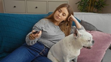 Genç sarışın bir kadın köpek evcil hayvanıyla oturma odasında dinleniyor. Uzaktan kumandayla evde sakin bir sahne gösteriyor..