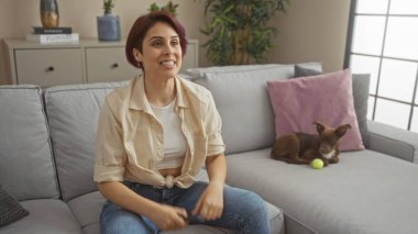 Kafkasyalı genç bir kadın rahat bir oturma odasında köpeğiyle birlikte kanepede dinleniyor..