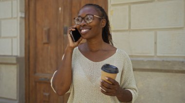 Afro-Amerikalı kadın elinde kahve fincanı tutarken telefonda konuşuyor.