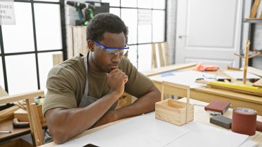 Atölyedeki düşünceli Afrikalı Amerikalı adam güvenlik gözlüğü takan marangozluk planlarını analiz ediyor..