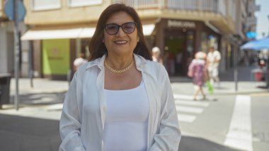 Güzel, orta yaşlı, kısa saçlı ve gözlüklü bir İspanyol kadın güneşli bir günde şehir sokaklarında kendine güvenerek gülümsüyor..