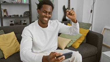 Modern bir oturma odasında akıllı telefonunu rahatça kullanan gülümseyen bir Afrikalı..
