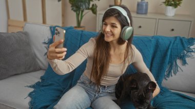 A young hispanic woman wearing headphones takes a selfie with her french bulldog while sitting on a couch in the living room of her apartment. clipart