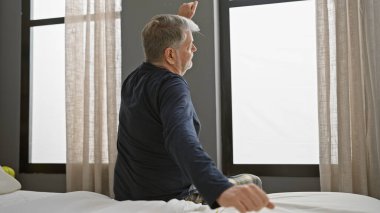 Parlak bir yatak odasında uyandıktan sonra yatakta gerinen gri saçlı bir adam..