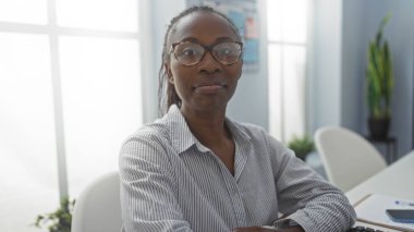 Ofis ortamında gözlük takan bir kadın, iş yerinde masa başında oturan Afrikalı Amerikalı genç bir kadın, kapalı alanda profesyonel bir ortam.