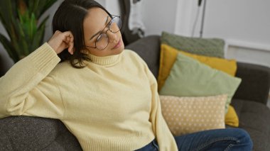 Genç İspanyol bir kadın gözlük takıyor. İçerideki bir kanepede, renkli yastıklarla, rahat rahat oturuyor.