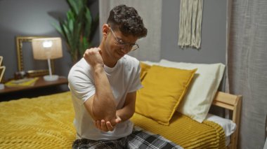 Genç İspanyol bir adam yatak odasında dirseğini acı dolu bir ifadeyle tutuyor sarı ve beyaz yastıklı düzgün bir yatakta oturuyor ve günlük giysiler giyiyor..