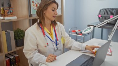 Genç, çekici, sarışın, beyaz bir kadın klinikte veteriner olarak çalışıyor, laptopuna odaklanmış, veteriner aletleri ve ekipmanlarıyla çevrili, profesyonel bir çalışma ortamını sergiliyor..