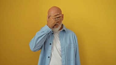 Gri saçlı ve sakallı bir adam mavi gömlekli, yüzü elle kaplı, canlı sarı bir arka planda duruyor.