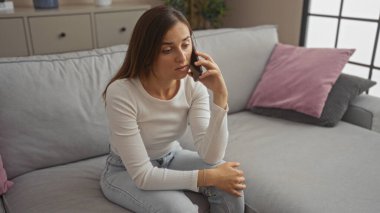 Beyaz bir kadın telefonda konuşuyor. Evinin oturma odasında oturuyor. Endişeli ve düşünceli görünüyor..