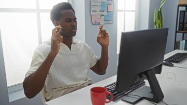 Genç, yakışıklı, Afro-Amerikan bir adam ofiste oturmuş bilgisayarın önünde parmaklarını çapraz yapıyor.