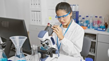 Laboratuvar ortamında mikroskop kullanan İspanyol kadın bilim adamı. Etrafı laboratuvar ekipmanlarıyla çevrili..