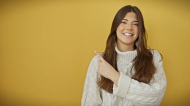 Sarı arka plan üzerinde beyaz kazak giyen gülümseyen genç İspanyol kadın reklam için boş bir alana işaret ediyor..