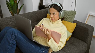 Günlük giysiler içinde genç bir İspanyol kadın evde çalışıyor, elinde dizüstü bilgisayar ve kulaklıkla kanepede oturuyor..